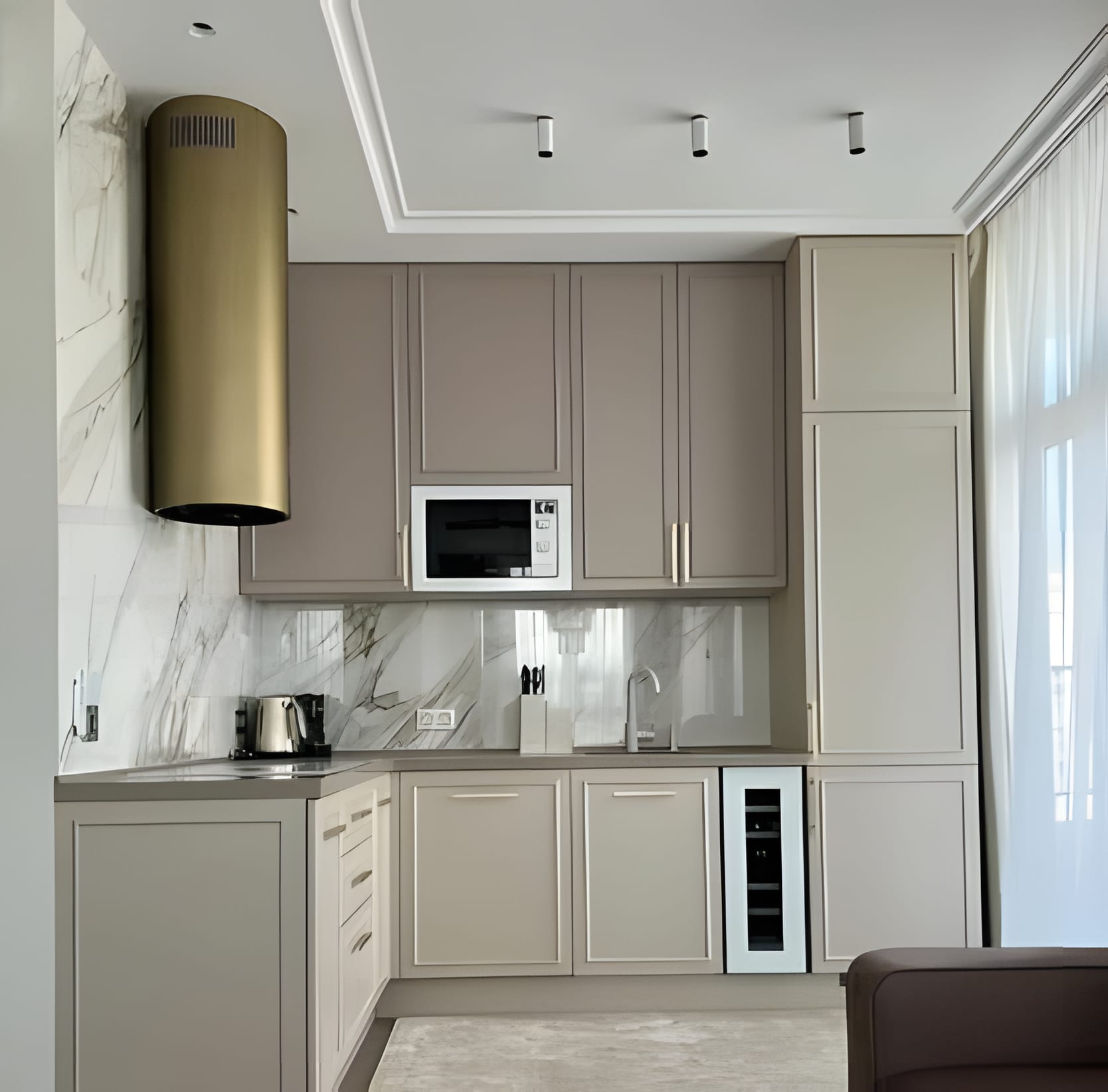 A pale L-shaped kitchen with soft-grey framed doors, a marble backsplash, and a brushed-brass cylinder hood as the room's one bold stroke.